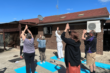 Group of NDIS participants in the Morning Mindfulness program, standing outside on yoga mats with hands clasped to the sky