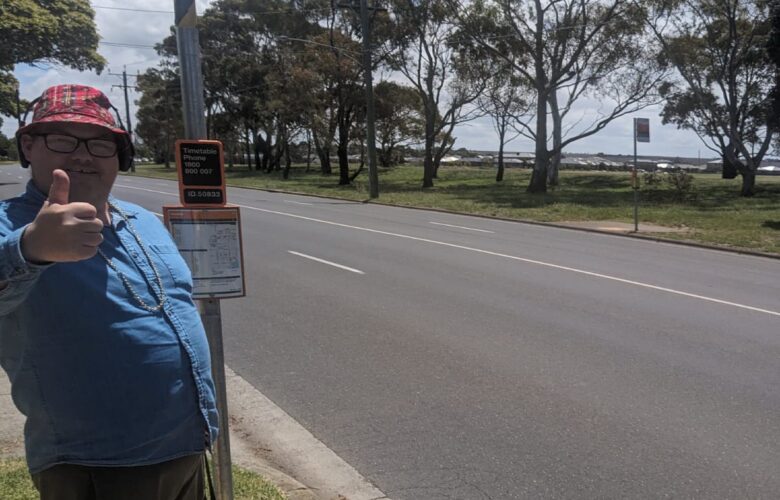 NDIS participant standing at bus stop on the side of the road giving thumbs up to camera