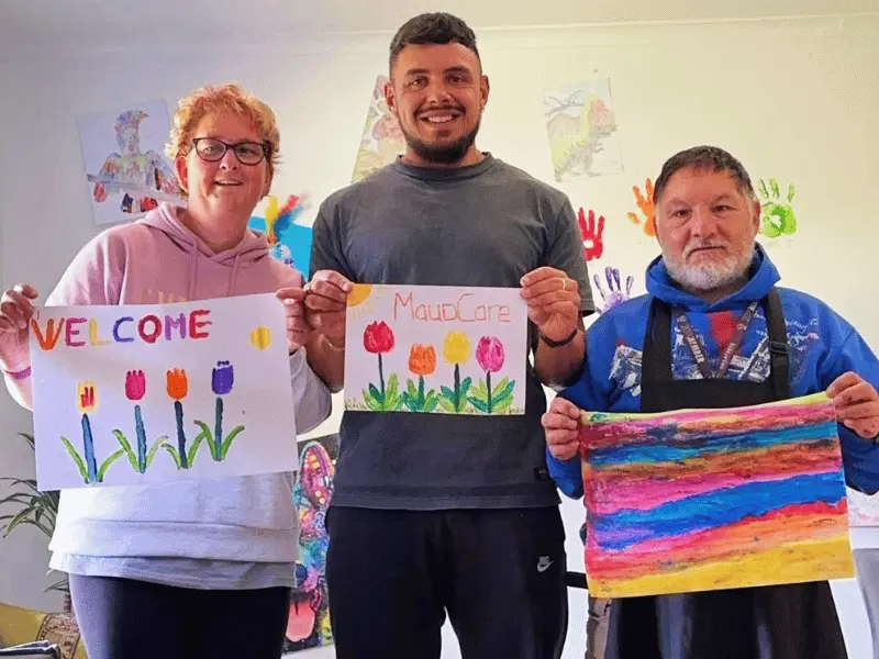 Two NDIS participants with support worker smiling at camera and each holding up their piece of artwork